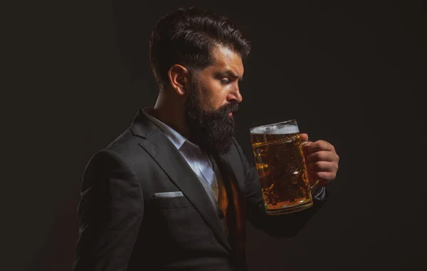 穿着经典西装的男人喝啤酒。身穿商务服装、满脸胡须的人看上去很高兴,也很满意.带着黑色背景的高杯啤酒的男人的画像. — 图库照片