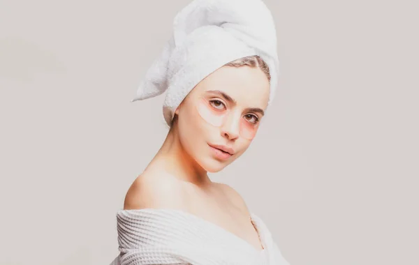 Ένα νεαρό όμορφο κορίτσι με μια πετσέτα στο κεφάλι και ένα λευκό κορίτσι ρόμπα φροντίζει το δέρμα χρησιμοποιώντας μπαλώματα κάτω από τα μάτια της. Εφαρμόζω μάσκα κατά της κόπωσης. Κορίτσι μειώσει σακούλες ματιών. — Φωτογραφία Αρχείου