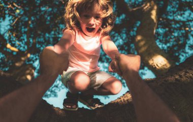 Baba oğlunun ağaca tırmanmasına yardım ediyor. Küçük çocuk ağaca tırmanmaya çalışıyor. Ebeveyn, çocuğun elini tutar..