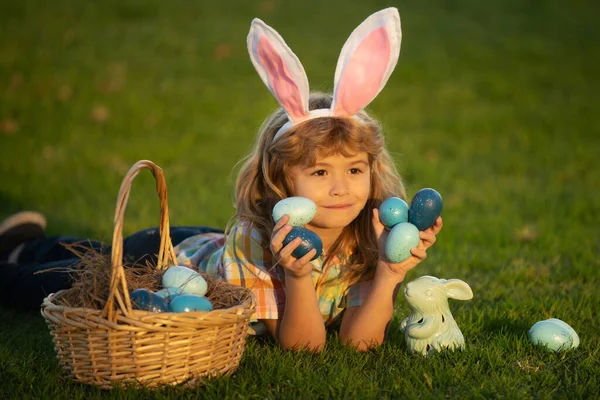 Mały króliczek z króliczymi uszami. Chłopiec z wielkanocnymi jajkami i koszykiem na trawie. Chłopiec polujący na wielkanocne jajka, leżący na trawie. Zajączek wielkanocny. — Zdjęcie stockowe
