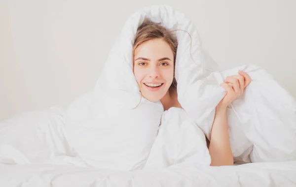 Mujer divertida con cara hermosa está sentada en la cama, envuelta en una manta suave. Mujer joven acostada en la cama y escondida debajo de la sábana. —  Fotos de Stock