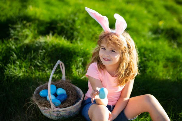 Konijnenpaaskind. Kinderen jagen op paaseieren. Jongen met paaseieren en konijnenoren in de achtertuin. Paasvakantie. — Stockfoto