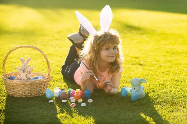 Pasen kinderen jongen in konijnenoren verf paaseieren buiten. Schattig kind in konijn kostuum met konijn oren hebben plezier in park schilderen eieren. — Stockfoto