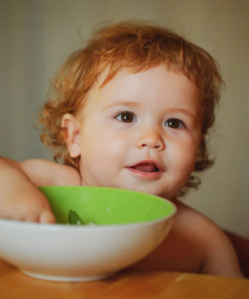 Portret van een grappig jongetje dat eet van een bord vol lepels. Grappig kind gezicht close-up. — Stockfoto
