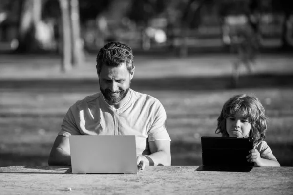 Uczenie się przez Internet. Ojciec i syn z laptopem studiujący online w zielonym parku letnim. Mały dzieciak siedzący przy stole i oglądający lekcje online na notatniku, tatuś pomagający dziecku odrabiać lekcje. — Zdjęcie stockowe