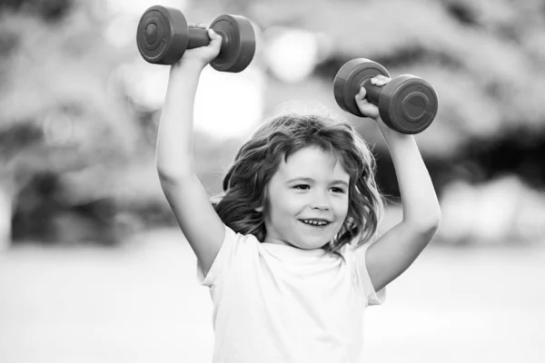 Happy little excited boy doing exercises with dumbbells in green park. Closeup portrait of sporty smiling child with dumbbells. Happy child boy exercising outdoor. — Foto Stock