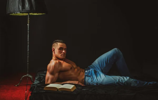 Homem sem camisa muscular na leitura do quarto. Macho nu na cama. Conceito de sexo e relaxamento. Amante do livro. — Fotografia de Stock