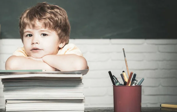 Μικρό αγόρι μαθητής με χαρούμενη έκφραση προσώπου κοντά στο γραφείο με σχολικά είδη. Το παιδί είναι έτοιμο να απαντήσει με έναν πίνακα σε φόντο. Ημέρα δασκάλων. — Φωτογραφία Αρχείου