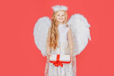 Hediyesi olan küçük bir melek çocuk. Beyaz kanatlı küçük melek hediyesini tutuyor. Neşeli melek gibi küçük kız. Meleklerin kanatlarıyla tatlı melek kız, kırmızıda izole edilmiş. Sevgililer Günü Kutlaması.