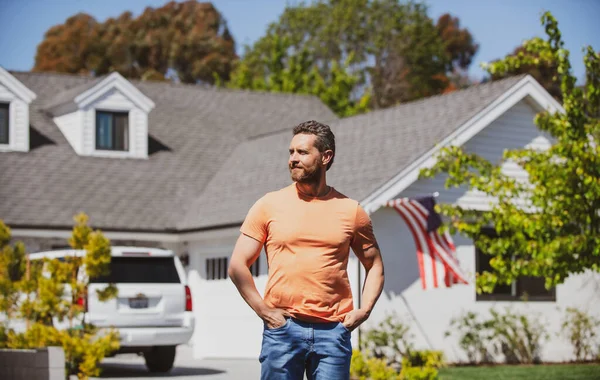 Retrato de um tipo a rir com as mãos nos bolsos encostadas à casa americana. Homem parado em frente à sua nova casa. Pessoas reais. — Fotografia de Stock