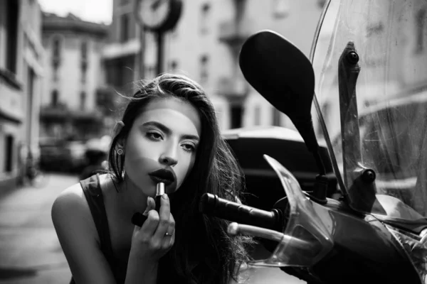 Makeupen i rörelse. Mode flicka med röda läppar sätta läppstift tittar i en spegel av motorcykel. Gatumodell. Stadsliv. — Stockfoto