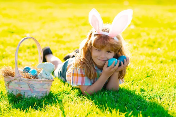 Een jongetje dat op paaseieren jaagt en op gras legt. Kind in konijn konijnenoren buiten. — Stockfoto