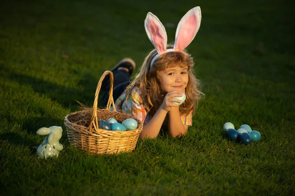 Tavşan kulaklı tavşan çocuklar. Paskalya yumurtalarını sepete koyan çocuk. Çocuk parkta çimlerin üzerinde yatıyor. Paskalya yumurtası avı. Fynny çocuk portresi. — Stok fotoğraf
