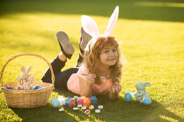 Kind jongen met paaseieren en konijnenoren leggen op gras schilderen eieren. — Stockfoto