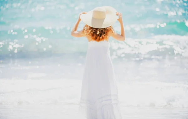 Вид сзади красивая девушка в соломенной шляпе, наслаждающаяся загоранием на пляже. Молодая загорелая женщина наслаждается бризом на море. Беззаботная женщина улыбается на заднем плане морским океаном. — стоковое фото