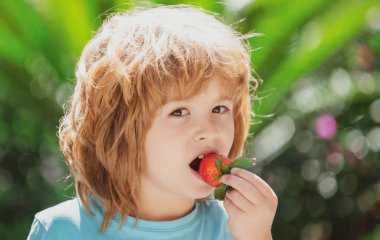 Sağlıklı çocuk yemekleri. Yeşil bahar arka planında çilek yiyen sevimli bir çocuk. Mutlu yüzlü çocukları kapatın..