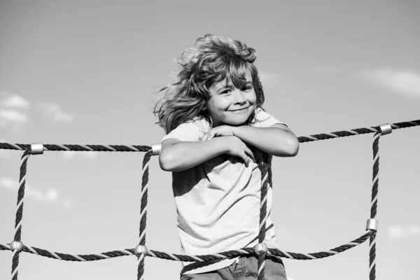 Gyönyörű mosolygós, aranyos fiú a játszótéren. Gyerekprogramok. A kölyök mászik a hálóra. Gyermeksport. — Stock Fotó