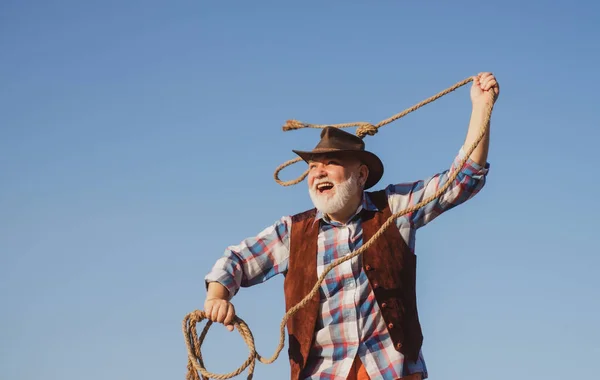 Senior western cowboy throwing lasso rope. Bearded wild west man with brown jacket and hat catching horse or cow. Rodeo or ranch. — Stock Photo, Image