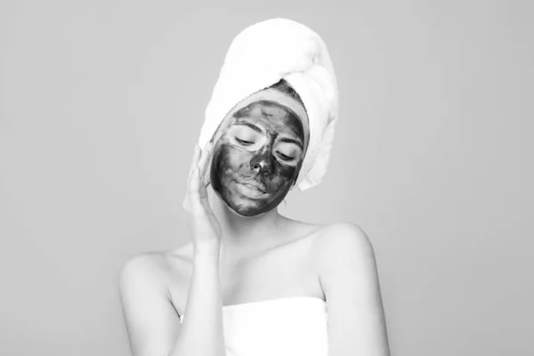 Vrouw met houtskoolmasker die haar neus aanraakt. Mud gezichtsmasker, gezichtsmasker spa. Mooie vrouw met cosmetische modder gezichtsbehandeling, spa gezondheid concept. Schoonheidsbehandeling voor de huid. Handdoek op het hoofd. — Stockfoto