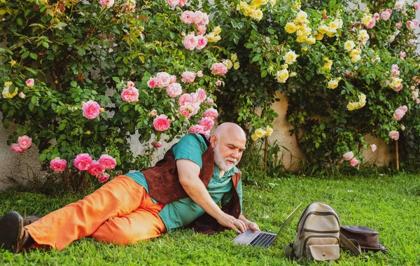 Anciano en el jardín de rosas con portátil en línea. Concepto de red social. Feliz vejez. — Foto de Stock