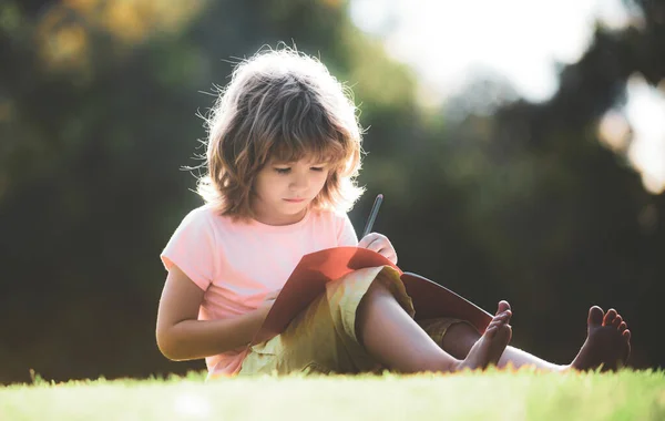 Забавный мальчик делает домашнюю работу и читает в парке на траве. Дистанционное образование для детей. Ученик дошкольного возраста. — стоковое фото