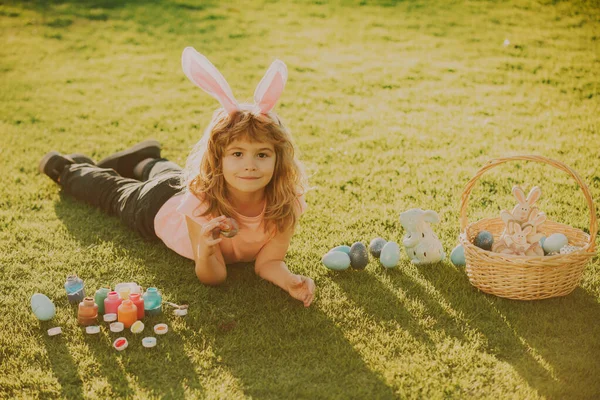 带着复活节彩蛋和兔子耳朵的孩子躺在草地上.复活节兔子耳朵和草地背景的孩子. — 图库照片