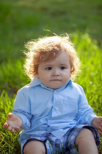 Retrato de lindo niño caucásico sentado en la hierba en el parque. Niño en césped de hierba verde durante el paseo en el patio. Feliz infancia y cuidado del bebé. —  Fotos de Stock