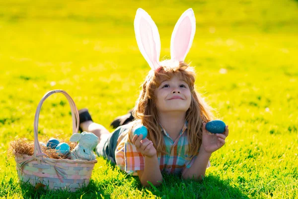 Kids jongen in konijnenoren jagen op paaseieren buiten. Een kind dat Pasen heeft, op gras ligt. Paaseieren jacht. — Stockfoto