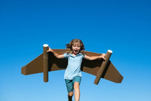 ジェットパックのスーパーヒーローと子供。夏の空を背景にした子供パイロット。紙飛行機のフライトを持つ男の子、段ボールの翼を持つおもちゃの飛行機、想像力、子供の自由. — ストック写真