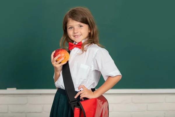 Primer plano retrato de pequeña y atractiva niña alegre sentada en el escritorio de la mesa en la sala de clase en el interior. Pequeña cara divertida de colegiala. Educación, aprendizaje y niños. —  Fotos de Stock