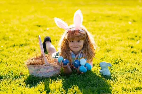 Весенние каникулы Мальчик лежит на траве и находит пасхальные яйца. Мальчик с пасхальными яйцами и кроличьими ушами на траве. — стоковое фото