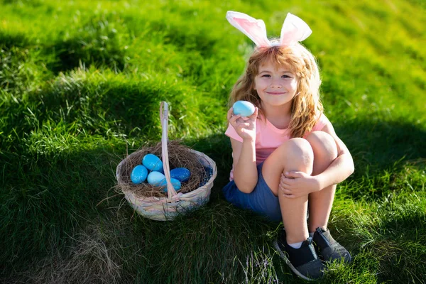 Feliz Pascua para los niños. Niño en orejas de conejo con huevos de colores jugar y la caza de huevos de Pascua fuera. Lindo niño con cesta de Pascua en la hierba. —  Fotos de Stock