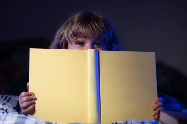 Rostro cercano del niño pequeño leyendo un libro en la casa oscura. Cara de niños con luz nocturna. —  Fotos de Stock
