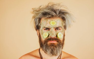 Yüzünde balçık maskesi ve salatalık dilimleri olan bir adam. Spa, cilt bakımı, sağlık ve yüz bakımı konsepti. Adamın kozmetik nemlendirici maskesi ve izole edilmiş bir stüdyo geçmişi var. Erkek yüz bakımı.