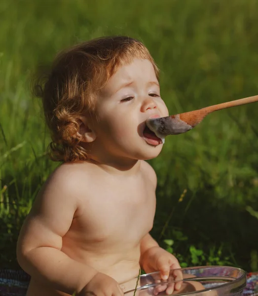 Colher alimentando bebê. Engraçado bebê no parque come-se com colher de madeira grande. Fundo de natureza turva. Alimentação saudável para crianças. Nutrição infantil. — Fotografia de Stock