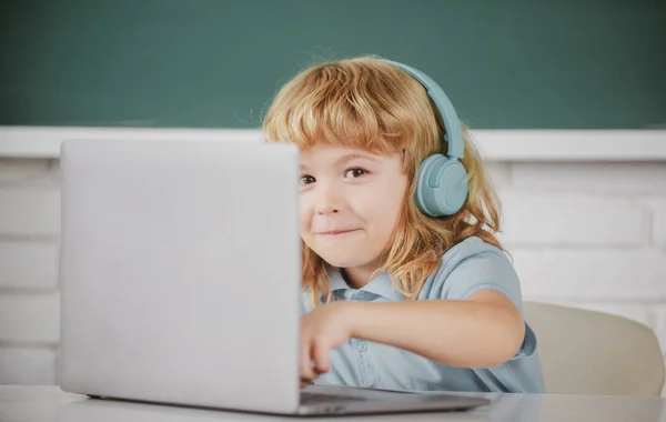 Primer plano retrato de niño pequeño en los auriculares ver video lección en la computadora en la escuela, niño en los auriculares tienen lección en línea, utilizando el ordenador portátil. — Foto de Stock