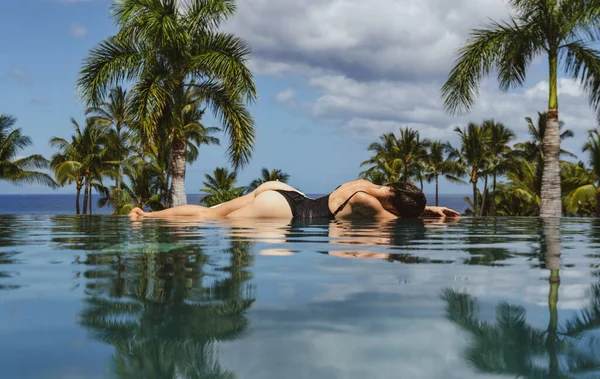 Luxury infinity pool. Young sexy woman enjoying the scenic ocean and tropical view from the edge of infinity pool on villa. Back view of a female in bikini at swimming pool.