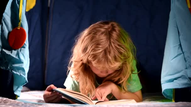 Niños leyendo un libro en la casa oscura. Niño leyendo un libro acostado en la cama. Niño sentado en la sala de estar viendo fotos en el libro de cuentos. Niño haciendo deberes para la escuela primaria. — Vídeo de stock