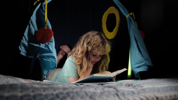 Kinder lesen im dunklen Haus ein Buch. Ein kleiner Junge liest ein Buch, das auf dem Bett liegt. Seriöses Kind liest Bücher, sieht ernst und konzentriert aus, spielt zu Hause, liest Geschichten oder Märchen. — Stockvideo