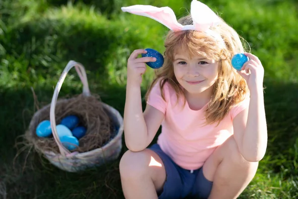 Paaskinderen. Konijnenpaaskind. Kinderen jagen op paaseieren. Jongen met paaseieren en konijnenoren in de achtertuin. Pasen mand — Stockfoto