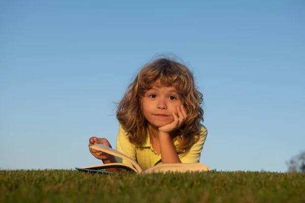 Niño divertido leyendo libro, tendido en la hierba en el campo en el fondo del cielo. Retrato de niños inteligentes. —  Fotos de Stock