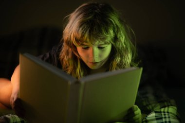 Küçük bir çocuk karanlık evde kitap okuyor. Çocukların yüzü gece lambasıyla kaplıdır. Eğitim ve okuma kavramı.