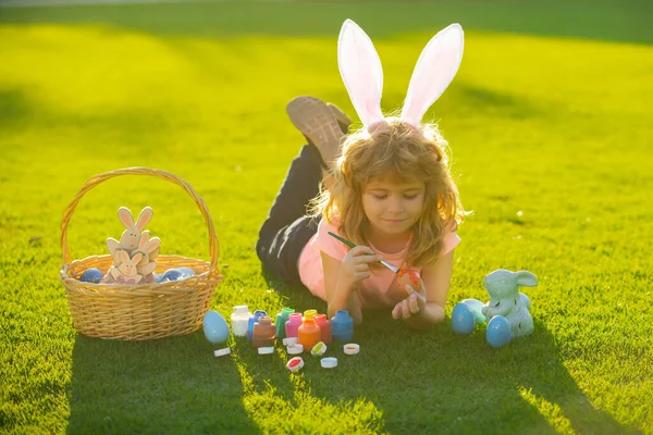 春天公园，身穿兔子服装、长着小兔子耳朵的小男孩在草地上画复活节彩蛋. — 图库照片