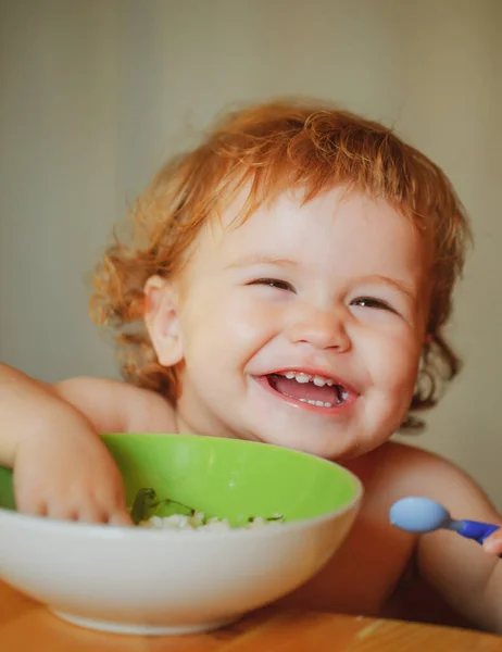Vtipné dítě, které jí jídlo se lžící v kuchyni. Usmívající se dítě jíst jídlo. — Stock fotografie