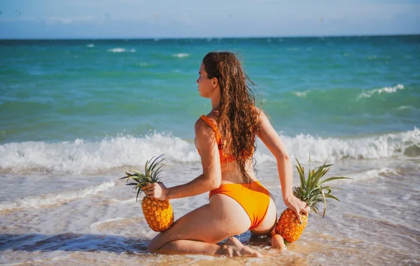 Beautiful caucasian sunbathed woman model in swimsuit posing on summer beach with white sand on blue sky and ocean background. Buttocks, butt, ass with pineapples. — Stockfoto