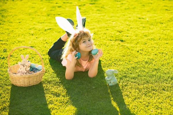 Feliz Pascua. Niño con traje de conejo con orejas de conejo cazando huevos de Pascua en la hierba en el parque de primavera. —  Fotos de Stock