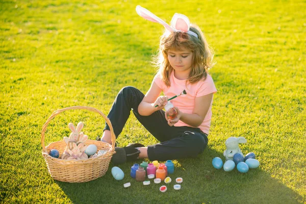 Пасхальный мальчик в кроличьих ушах рисует пасхальные яйца на открытом воздухе. Симпатичный ребенок в кроличьем костюме с кроличьими ушами, веселящимися в парке. — стоковое фото