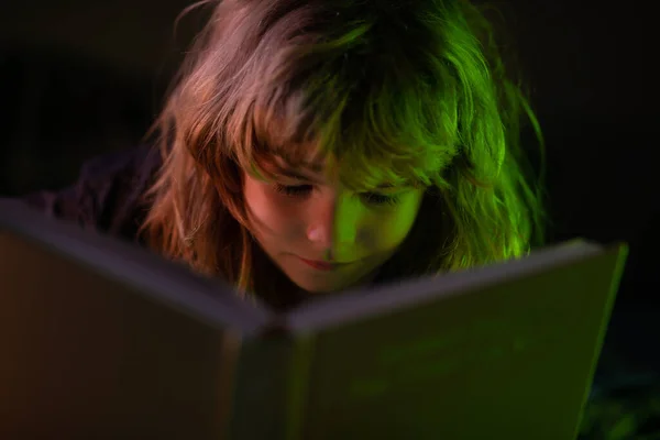 Ребенок читает сказки на ночь в темной комнате. Маленький милый мальчик, читающий книгу, лежащую на кровати. Ребёнок читает книгу на ночь перед сном. — стоковое фото