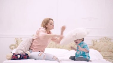 Mutlu bir aile çocuğu ve anne yataktaki yatakta zıplıyor neşeli bir anne yatak odasında sevimli bir çocukla eğlenceli bir oyun oynuyor. Anne ve çocuk yatak odasında yastıkla oynuyorlar..