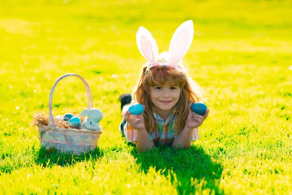 Niño cazando huevos de Pascua. Niño acostado en la hierba y encontrando huevos de Pascua. Niño con huevos de Pascua y orejas de conejo en la hierba. — Foto de Stock